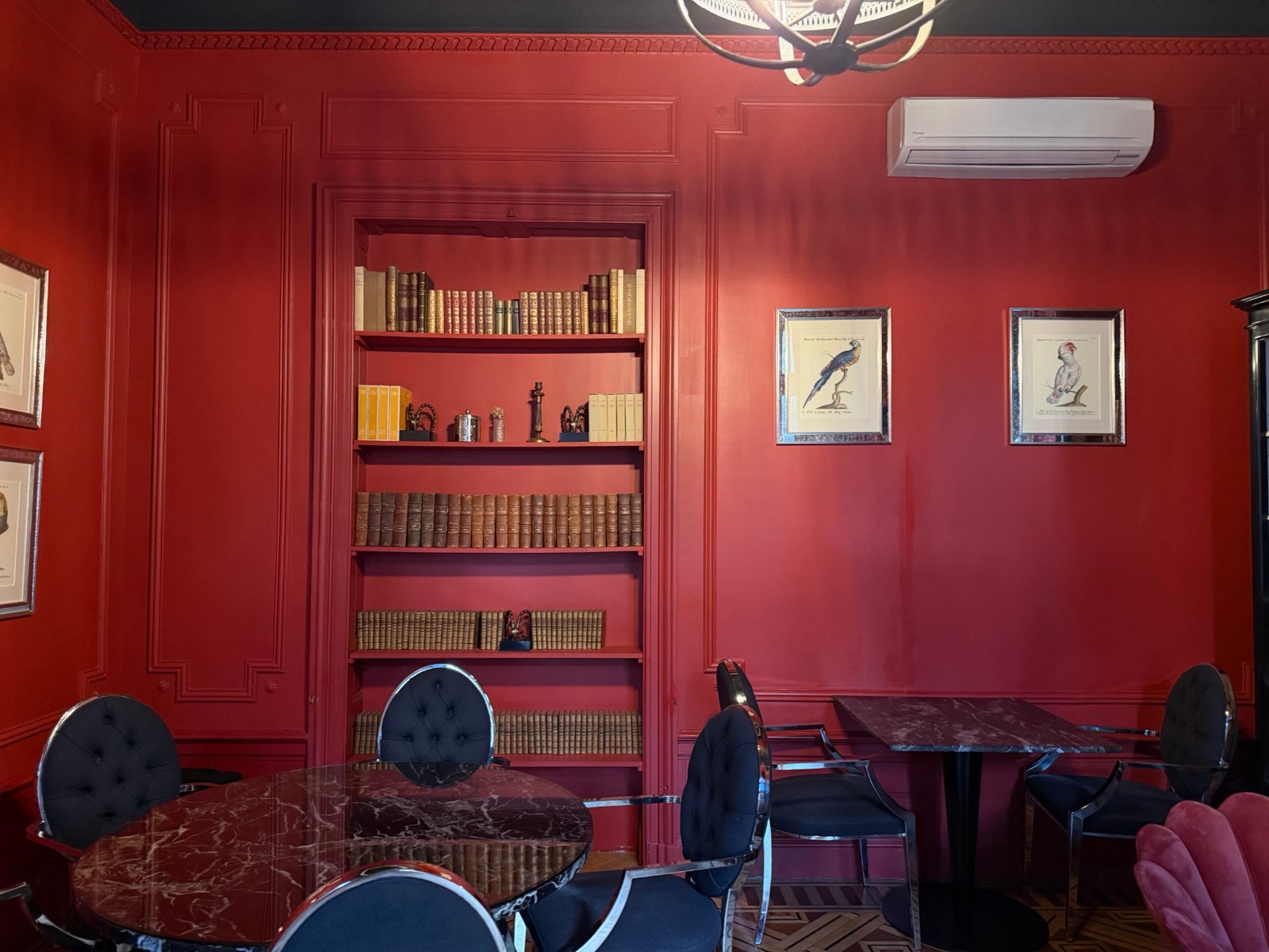 Salle rouge du Palais Gallien avec bibliothèque encastrée, gravures de perroquets, tables en marbre sombre et fauteuils bleu nuit